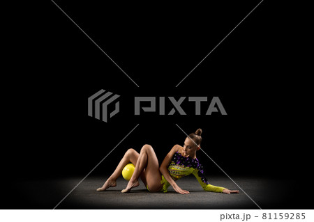 Portrait of little girl, rhythmic gymnastics artist in bright stage costume isolated on dark studio background in ray of light. Concept of sport, action, aspiration, active lifestyle 81159285