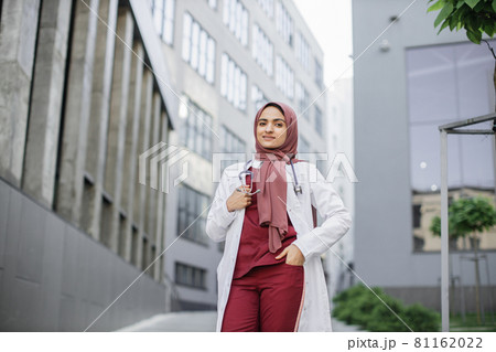 Beautiful Malay lady doctor wearing hijab, eyeglasses and stethoscope outside modern clinic 81162022