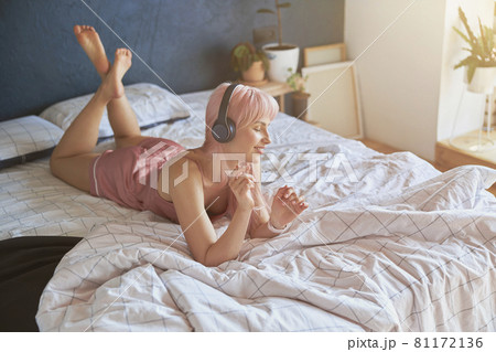 Stylish woman wearing pink silk pajamas enjoys with headphones lying on comfortable bed 81172136