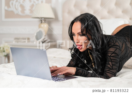 A sexy brunette with big breasts dressed in black underwear lies on the bed and flirts over video communication on a laptop. The woman works as a web model 81193322