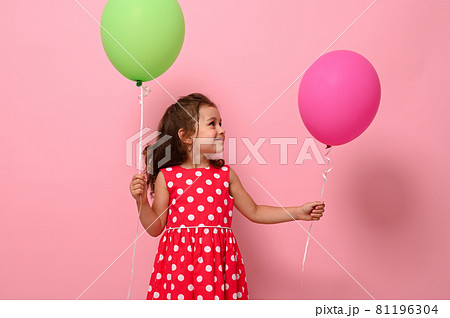 Beautiful gorgeous 4 year pretty birthday girl in pink polka dot dress, holds two multicolored balloons, rejoices looking at a green balloon in her hand, isolated on pink background with copy space. 81196304