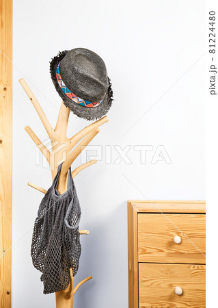Minimalistic hallway interior with trendy wooden clothes hanger stand with gray staw hat and mesh bag, and natural wooden commode against a gray wall. Copy space 81224480