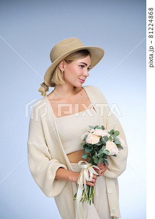 A young beautiful blonde caucasian woman with a short haircut in a beige suit and hat with bouquet of white flowers on blue background. Beauty portrait. The girl smiles, clean skin and white teeth. 81224868