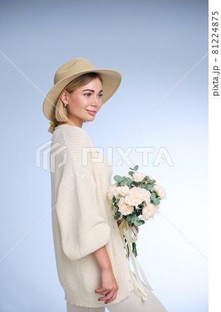 A young beautiful blonde caucasian woman with a short haircut in a beige suit and hat with bouquet of white flowers on blue background. Beauty portrait. The girl smiles, clean skin and white teeth. 81224875