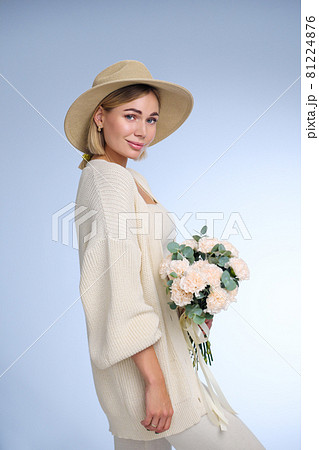 A young beautiful blonde caucasian woman with a short haircut in a beige suit and hat with bouquet of white flowers on blue background. Beauty portrait. The girl smiles, clean skin and white teeth. 81224876