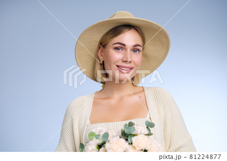 A young beautiful blonde caucasian woman with a short haircut in a beige suit and hat with bouquet of white flowers on blue background. Beauty portrait. The girl smiles, clean skin and white teeth. 81224877