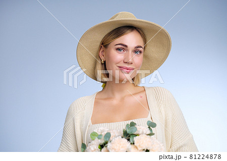 A young beautiful blonde caucasian woman with a short haircut in a beige suit and hat with bouquet of white flowers on blue background. Beauty portrait. The girl smiles, clean skin and white teeth. 81224878