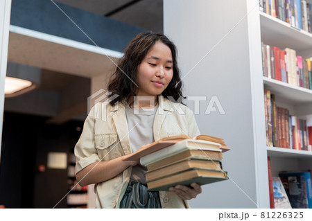 Happy girl with stack of books reading one of them 81226325