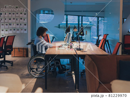 Disabled businessman in wheelchair working overtime alone at his desk in an office late at night Disabled businessman in wheelchair working overtime alone at his desk in an office late at night 81229970