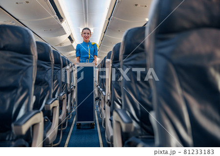 Female cabin attendant leading trolley cart through empty plane aisle 81233183