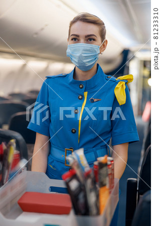 Stewardess in protective face mask looking away, serving food to passengers on aircraft. Air hostess walking with trolley on aisle 81233310