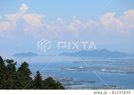 高台からの緑の樹々と琵琶湖大橋と沖島のある青い琵琶湖と田畑や街や霞む遠くの山やもくもくした雲と青空 81235830