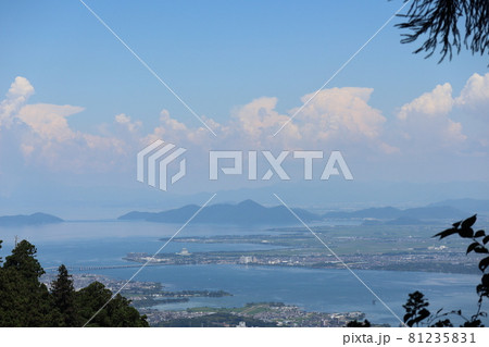 高台からの緑の樹と琵琶湖大橋と沖島のある青い琵琶湖と田畑や街や霞む遠くの山や町ともくもくした雲と青空 81235831