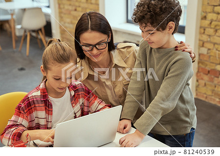 Experiment. Caring young female teacher in glasses smiling while helping smart kids to learn programming using laptop during STEM class 81240539