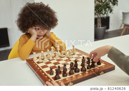 Cute player. Funny little boy with afro hair watching his friend making a move while playing chess, sitting at the table indoors 81240626