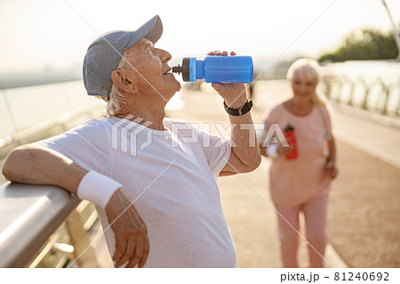 Positive senior man drinks water leaning onto railing while lady runs along footbridge Positive senior man drinks water leaning onto railing while lady runs along footbridge 81240692