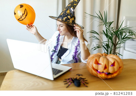 Halloween Witch hat.Woman with Pumpkin and laptop. Indoor portrait of cute young witch at home. Pretty young woman dressed as a fairy with pumpkin. Helloween people concept. 81241203