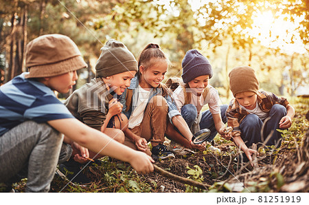 Happy school children boys and girls with backpacks making bonfire with magnifying glass in forest 81251919