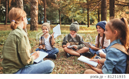 School children with notebooks and female teacher sitting on ground in forest during ecology lesson 81251935