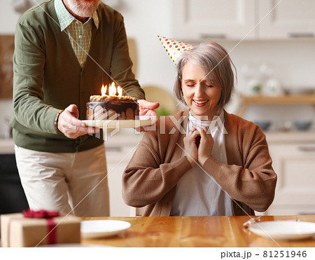 Happy elderly woman in party hat closing her eyes, she makes a wish, while her husband holds a chocolate cake with candles   during Birthday celebration 81251946