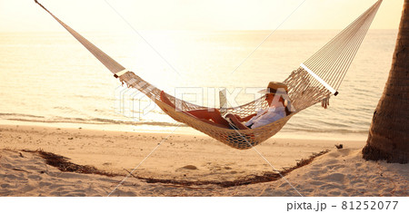 Young woman female freelancer using laptop while lying in hammock on the tropical beach at sunset 81252077
