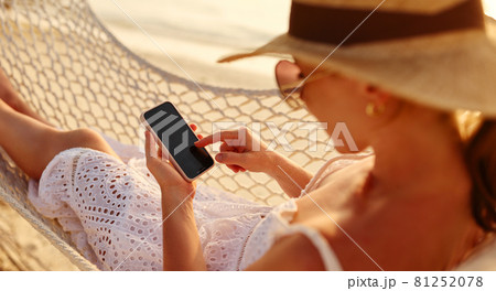 Working at sunset. Young happy female using smartphone while lying in the hammock on the beach Working at sunset. Young happy female using smartphone while lying in the hammock on the beach 81252078