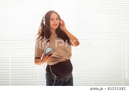 Portrait of pregnant woman near window at home and listening music in headphones. Pregnancy and leisure concept. Portrait of pregnant woman near window at home and listening music in headphones. Pregnancy and leisure concept. 81253382