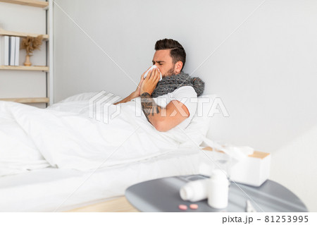 Home treatment for colds, flu and covid-19. Sick young guy blowing his nose in napkin, lying in bed Home treatment for colds, flu and covid-19. Sick young guy blowing his nose in napkin, lying in bed 81253995