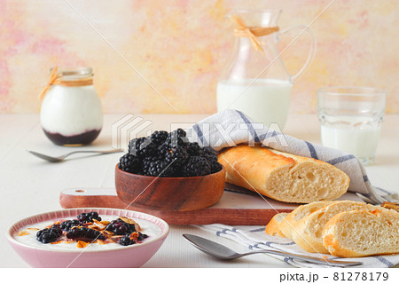 Bowl of blackberries, milk, yoghurt with oatmeals and bread, a table set for breakfast, ingredients from the local farmers market. Closeup, front view. 81278179