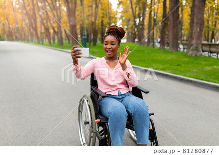 Happy young black disabled lady in wheelchair having video call on smartphone, waving at camera in autumn park 81280877