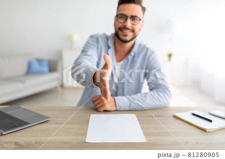 Job interview concept. Positive millennial man stretching hand for handshake, sitting at workplace, selective focus 81280905