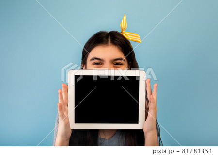 Indian teen girl hiding behind tablet computer with empty screen on blue background, mockup for website or app 81281351