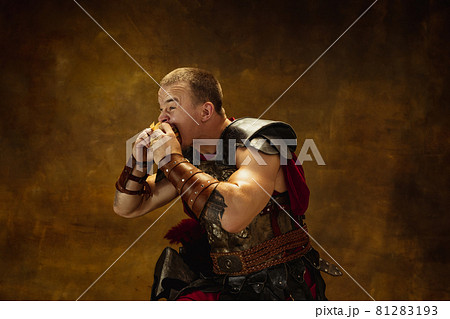 Portrait of young man, medieval warrior or knight in war equipment eating burger isolated on vintage dark background. Comparison of eras, art, history, humor, retro style concept Portrait of young man, medieval warrior or knight in war equipment eating burger isolated on vintage dark background. Comparison of eras, art, history, humor, retro style concept 81283193