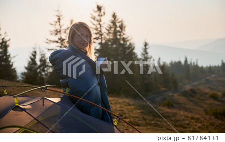 Portrait of blonde woman with nice smile on her face standing on mountain meadow near tent, enjoying taste of fragrant coffee on bright sunny morning. 81284131