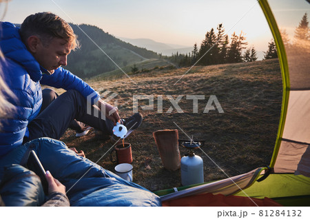 Handsome man traveler sitting on grassy hill near camp tent and pouring hot drink into cup. Male hiker making coffee while woman sitting in tourist tent and holding smartphone. Concept of camping. 81284132