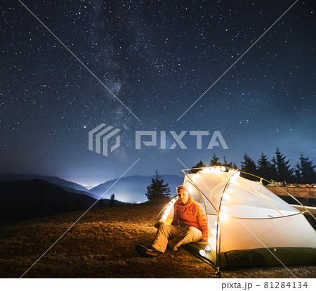 Young traveler sitting in his white tent, lit by light bulbs, on grass under open starry sky with Milky way. Night mountain camping against backdrop of mountain hills and light of city between them. Young traveler sitting in his white tent, lit by light bulbs, on grass under open starry sky with Milky way. Night mountain camping against backdrop of mountain hills and light of city between them. 81284134