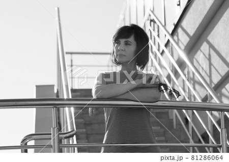 A brunette in a striped dress smiling stands on the stairs leaning her elbows on the railing and holding glasses in her hands 81286064