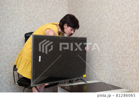 A brunette woman in the apartment installs a new large LCD TV on the table A brunette woman in the apartment installs a new large LCD TV on the table 81286065