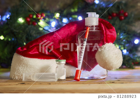 Covid-19 and Christmas holidays. Antiseptic hand sanitizer bottle, syringe and medicines on wood. Santa cap, coronavirus vaccines and syringe over Xmas tree Covid-19 and Christmas holidays. Antiseptic hand sanitizer bottle, syringe and medicines on wood. Santa cap, coronavirus vaccines and syringe over Xmas tree 81288923