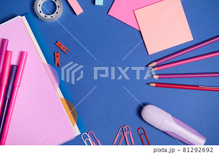 Assortment of school office accessories in pink shades, isolated on blue background, copy space. Copybook, felt tip pens, markers, paper clips,pencils and stationery supplies scattered in a circle 81292692
