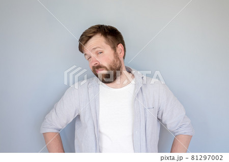 Caucasian bearded man in a shirt looks suspiciously and slyly, narrowing his eyes and akimbo. Gray 81297002
