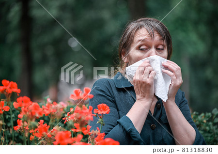 A woman blows her nose in a handkerchief in the garden with flowers in the foreground. Spring allergy concept. 81318830