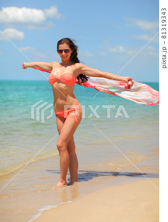 Young fit woman in bikini and sunglasses holding scarf waving behind her, calm sea in background. Young fit woman in bikini and sunglasses holding scarf waving behind her, calm sea in background. 81319343