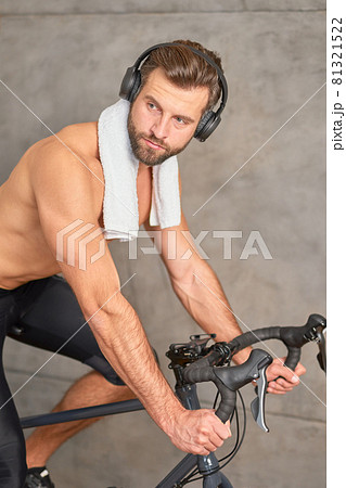 Bearded young man in headphones riding stationery bike Bearded young man in headphones riding stationery bike 81321522