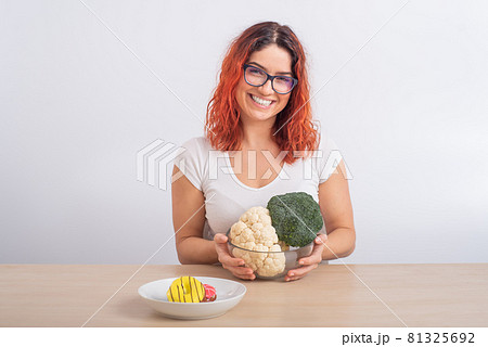 Caucasian woman prefers healthy food. Redhead girl chooses between broccoli and donuts on white background. Caucasian woman prefers healthy food. Redhead girl chooses between broccoli and donuts on white background. 81325692