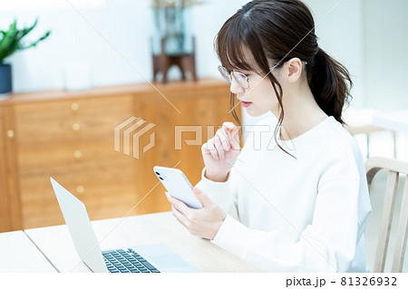 リビングでノートパソコンを使いながらスマホを操作する眼鏡の女性 81326932