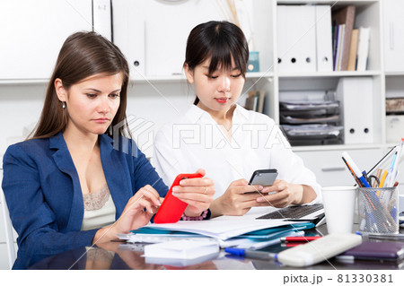 Two women write sms by phone in the office Two women write sms by phone in the office 81330381