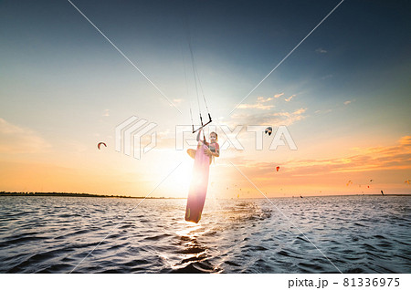 Young caucasian woman kitesurfer doing a jump trick on a sunset background. The girl hides the setting sun in a jump. double-sided copy space. Central composition. Kitesurfer spot 81336975