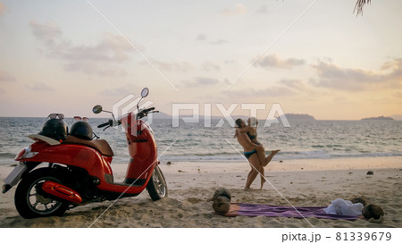 Scooter road trip. Lovely couple on red motorbike in white clothes on sand beach. People walking near the tropical palm trees, sea. Motorcycle rent. Scooter road trip. Lovely couple on red motorbike in white clothes on sand beach. People walking near the tropical palm trees, sea. Motorcycle rent. 81339679