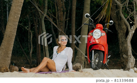 Scooter road trip. Woman alone on red motorbike in white clothes on sand beach by ocean. One girl caucasian tourist walk near tropical palm tree. Scooter road trip. Woman alone on red motorbike in white clothes on sand beach by ocean. One girl caucasian tourist walk near tropical palm tree. 81339688